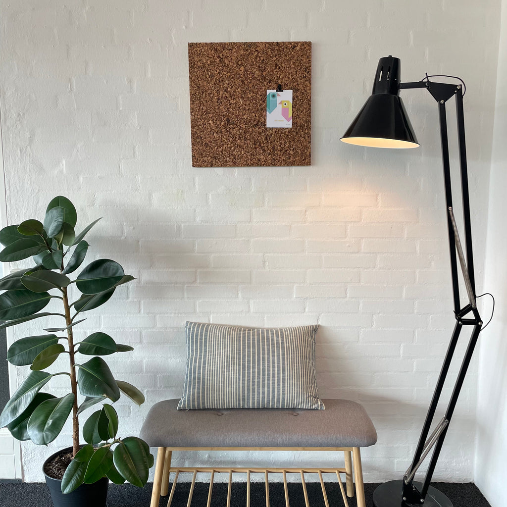 Rustik korktavle på væg, bænk med pude under, plante ved siden af, sort gulvlampe til højre.