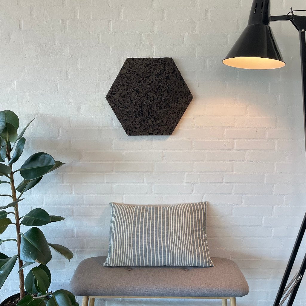 Mørk sekskantet korkpanel på hvid murstensvæg over bænk, ved siden af gulvlampe.