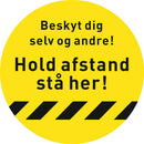 Gul cirkulær gulvsticker med sort tekst: "Beskyt dig selv og andre! Hold afstand stå her!"