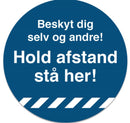 Blå cirkel med teksten "Beskyt dig selv og andre! Hold afstand stå her!" i hvid skrift.